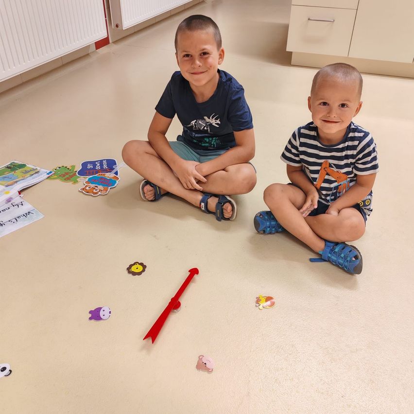 Zwei junge Jungen sitzen auf dem Boden, lächeln und spielen mit Spielzeug und einem roten Pfeil. Der Raum hat weiße Wände und einen weißen Boden.