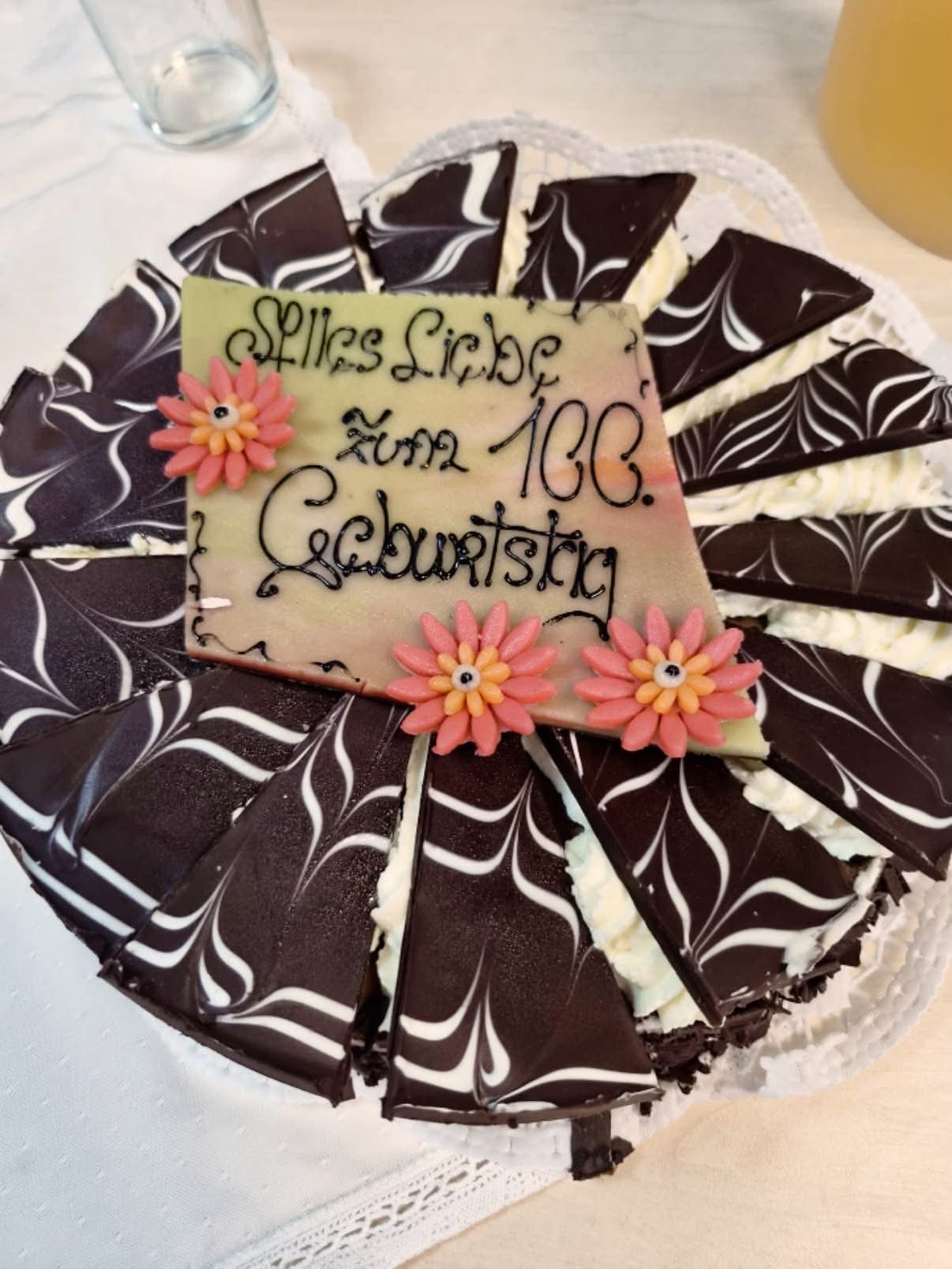 Ein Schokoladenkuchen mit einem rechteckigen Schild oben, das 'Spass Liebe Zum 100. Geburtstag' und zwei rosa Blumen zeigt. Der Kuchen ist in mehrere Stücke geschnitten.