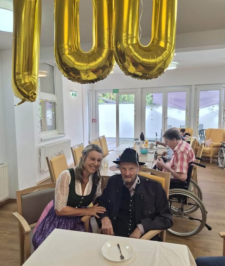 Ein älterer Mann und eine Frau sitzen lächelnd in einem Essbereich. Über ihnen hängt ein goldener Ballon mit der Nummer 100. Dahinter sitzt eine Person im Rollstuhl am Tisch.
