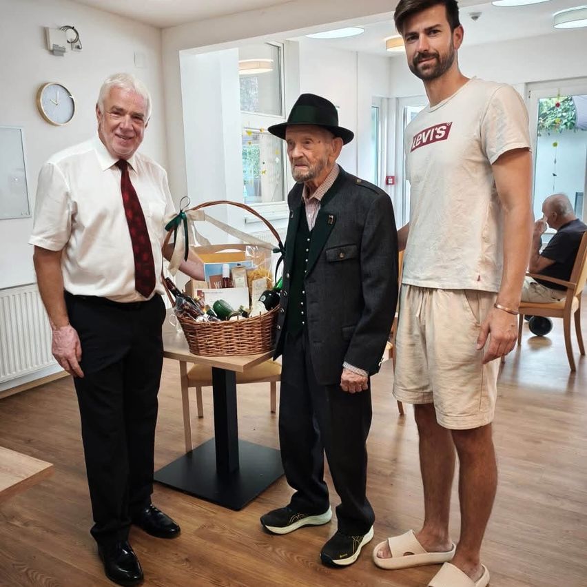 Drei Männer stehen in einem Raum, einer hält einen Korb mit Geschenken. Der Mann auf der linken Seite trägt eine Krawatte und Lederschuhe. Der Mann in der Mitte trägt einen Hut und Lederschuhe. Der Mann auf der rechten Seite trägt ein Levis-Shirt und Sandalen.