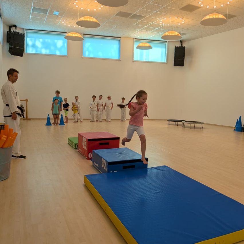 Ein kleines Mädchen springt auf einer blauen Matte in einem Fitnessstudio mit anderen Kindern und einem Trainer.