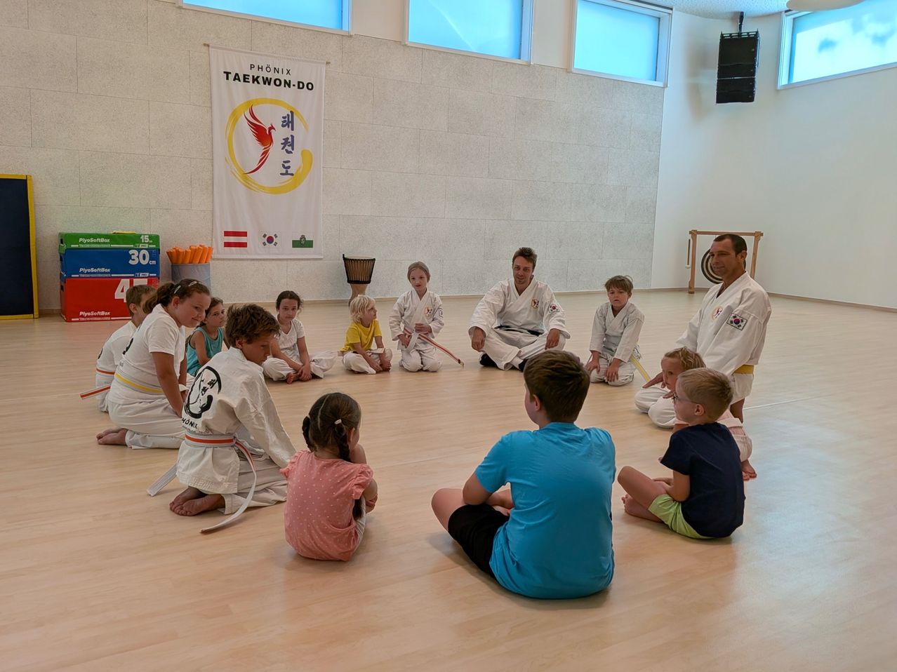Eine Gruppe von Kindern und Erwachsenen in Kampfsportuniformen sitzt im Kreis auf einem Holzboden, mit zwei Ausbildern in der Mitte. Ein Banner mit koreanischem Text hängt an der Wand.