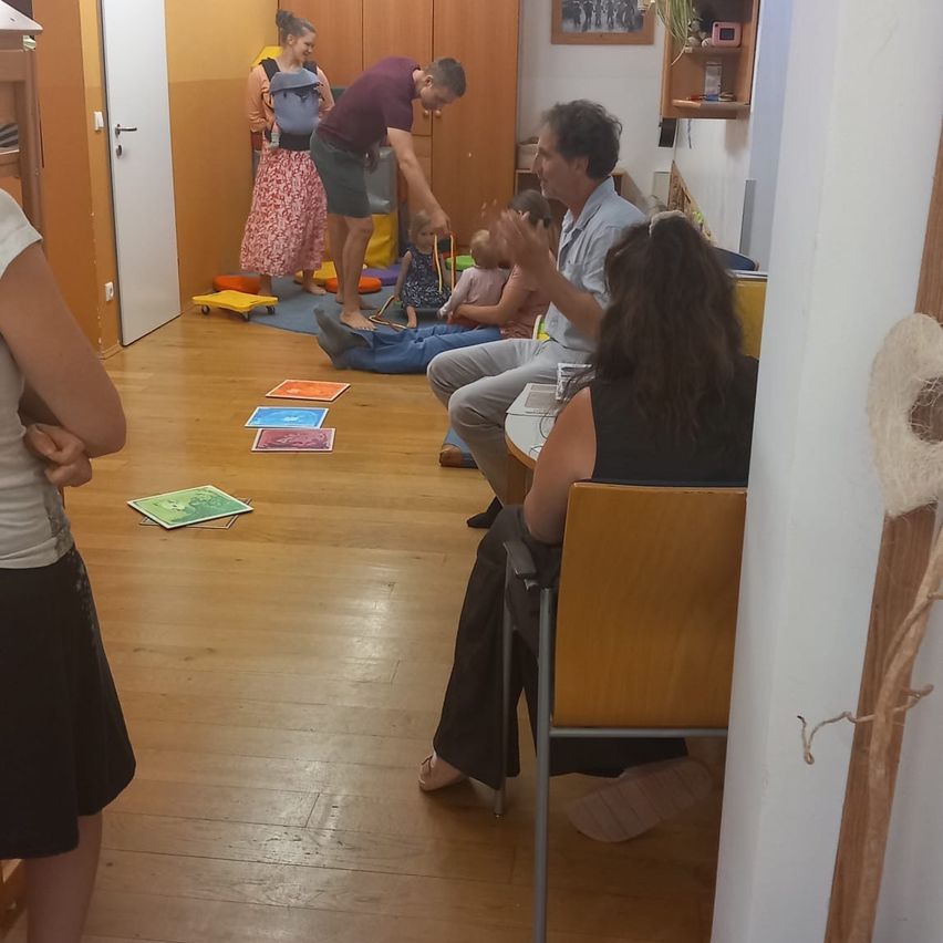 Ein Mann und eine Frau spielen mit einem Baby auf dem Boden. Ein Mann sitzt, während eine Frau in der Nähe der Tür steht. An der Wand hängt ein Bilderrahmen.