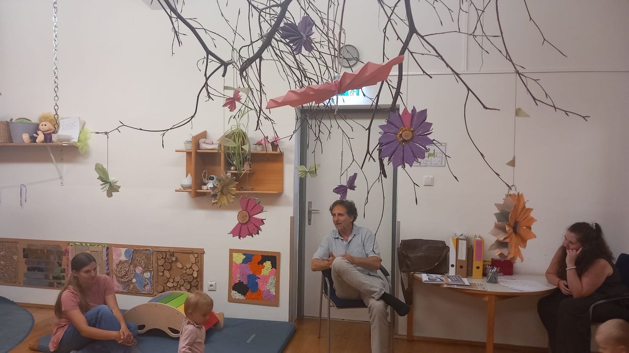 Ein erwachsener Mann sitzt in einem Stuhl in einem Raum mit einem Baby. Über ihm ist ein Baumzweig mit Blumen. Eine Uhr und eine Steckdose befinden sich an der Wand.