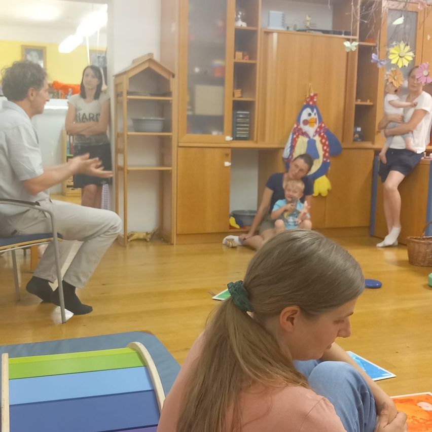 Ein Mann spricht zu einer sitzenden Frau, beide schauen ein Baby an. Eine andere Frau hält ein Baby, während ein Mann auf dem Boden sitzt. Sie sind in einem Raum mit Holzböden und Regalen.
