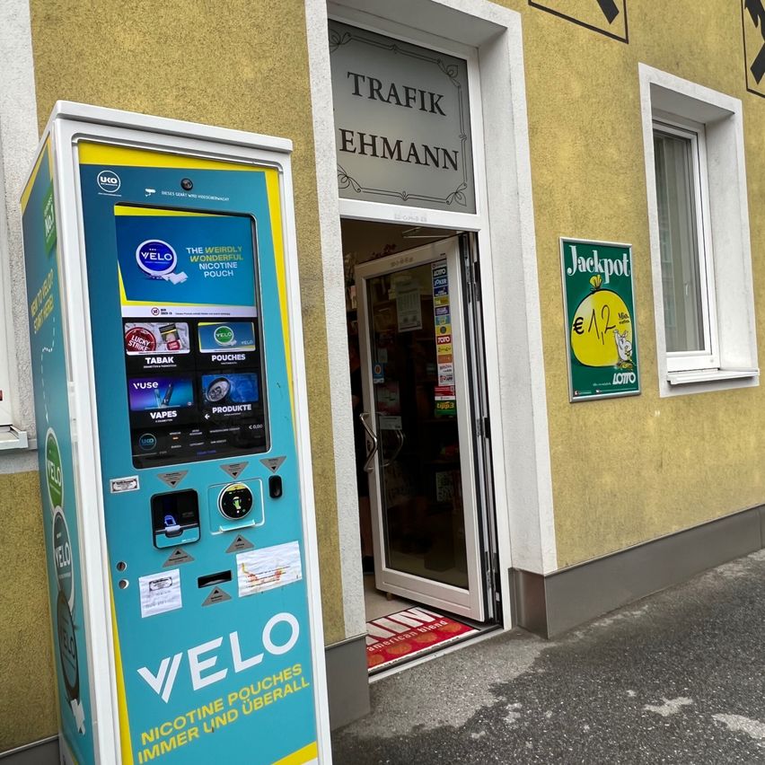 Ein blauer und gelber Velo-Automat steht vor einem gelben Gebäude mit einem Schild, auf dem Trafik Ehmann steht. Ein grünes Schild mit Jackpot und 1,12€ ist an der Wand.
