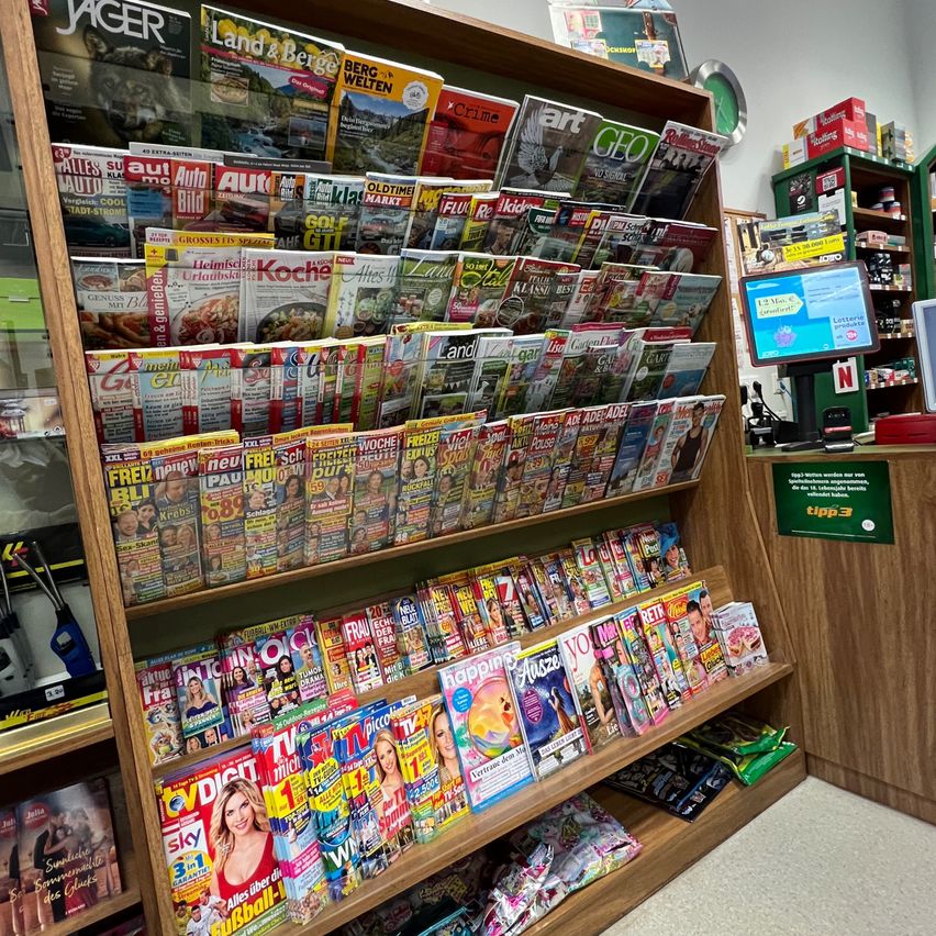 Ein Laden mit einem hölzernen Regal voller verschiedener Zeitschriften. Ein Monitor ist auf dem Tresen mit einem Schild platziert. Ein Regal mit Büchern befindet sich rechts.