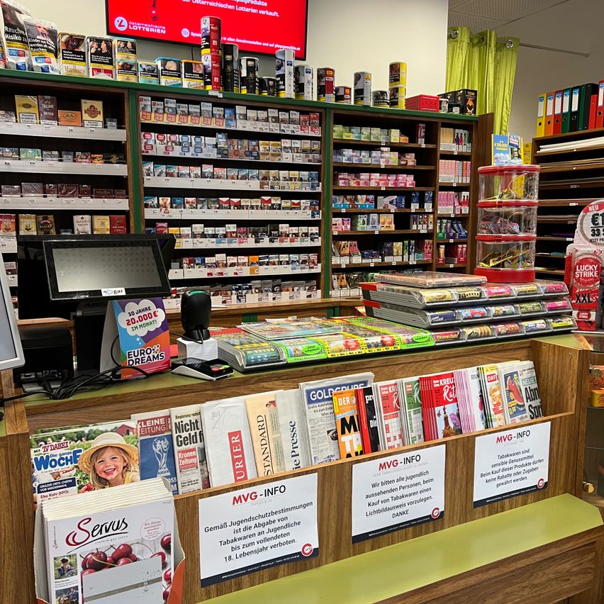 Ein Geschäft mit einer großen Auswahl an Produkten, einschließlich Zigaretten, Büchern und Zeitschriften. Ein Computermonitor und ein Schild für MGV-INFO befinden sich auf dem Tresen.