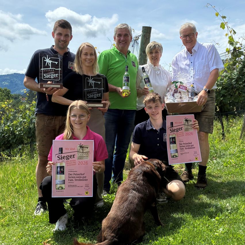 Eine Gruppe von Menschen in Grün, Pink und Schwarz posiert für ein Foto im Weinberg, hält Auszeichnungen und Weinflaschen in den Händen. Ein Hund sitzt neben ihnen auf dem Boden.