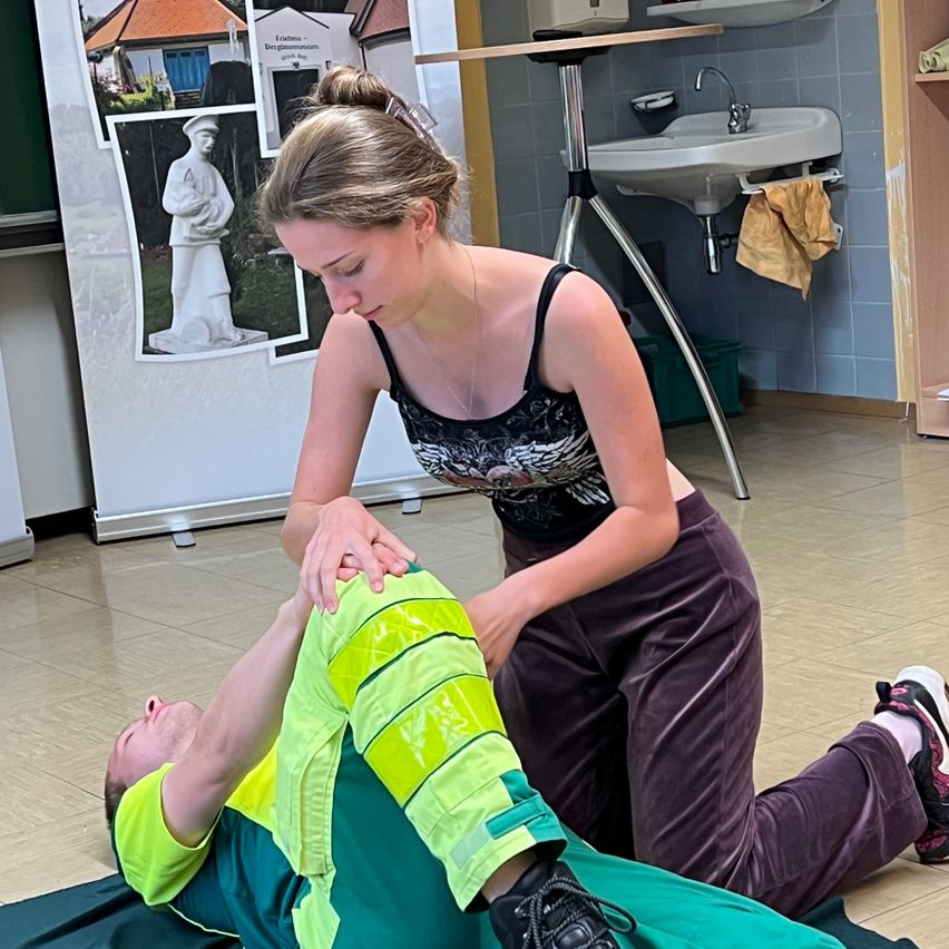 Eine Frau in einem schwarzen Tanktop und braunen Hosen führt eine medizinische Prozedur an einem Mann durch, der auf dem Boden liegt und eine Sicherheitsweste trägt. Hinter ihnen befindet sich ein Waschbecken mit einem Handtuch und ein Poster an der Wand.