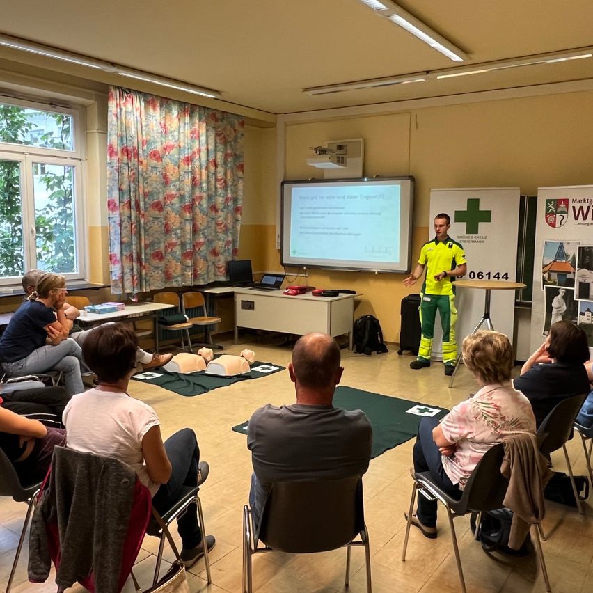 In einem Klassenzimmer steht ein Mann in gelber und grüner Uniform vor einer Projektionsleinwand und präsentiert einer sitzenden Zuhörerschaft. Übungspuppen und medizinische Geräte liegen auf dem Boden.