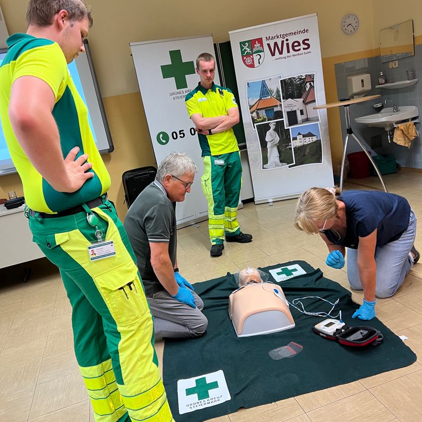 Erste-Hilfe-Training in vollem Gange. Mehrere Personen arbeiten an einem Mannequin. Eine Person kniet, eine andere sitzt, und zwei weitere beobachten. Ein Banner mit Text und einem Kreuz im Hintergrund.
