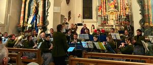 Eine Gruppe von Musikern spielt verschiedene Instrumente in einer Kirchenumgebung. Die Musiker umfassen Trompeten, Tubas und andere Blechblasinstrumente. Das Kircheninnere zeichnet sich durch komplizierte Designs und ein Weihnachtsthema aus.