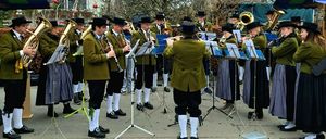 Eine Collage von Musikern, die im Freien spielen, in traditioneller Kleidung gekleidet und verschiedene Instrumente wie Trompeten und Xylophon halten. Die Musiker sind in Reihen angeordnet, einige stehen und andere sitzen. Der Hintergrund zeigt Bäume und ein Gebäude.
