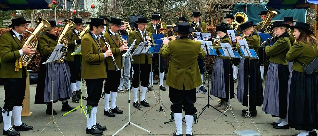Eine Collage von Musikern, die im Freien spielen, in traditioneller Kleidung gekleidet und verschiedene Instrumente wie Trompeten und Xylophon halten. Die Musiker sind in Reihen angeordnet, einige stehen und andere sitzen. Der Hintergrund zeigt Bäume und ein Gebäude.