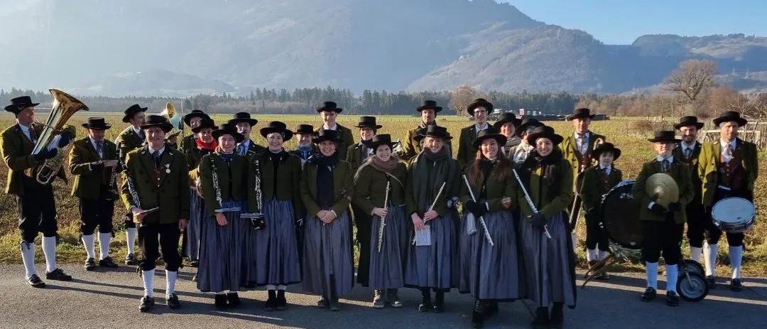 Zwei Fotos einer Marschkapelle in traditionellen Kostümen. Das erste Bild zeigt sie vor Bergen, das zweite mit einem Haus und einer Straßenlaterne im Hintergrund.