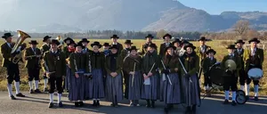 Zwei Fotos einer Marschkapelle in traditionellen Kostümen. Das erste Bild zeigt sie vor Bergen, das zweite mit einem Haus und einer Straßenlaterne im Hintergrund.