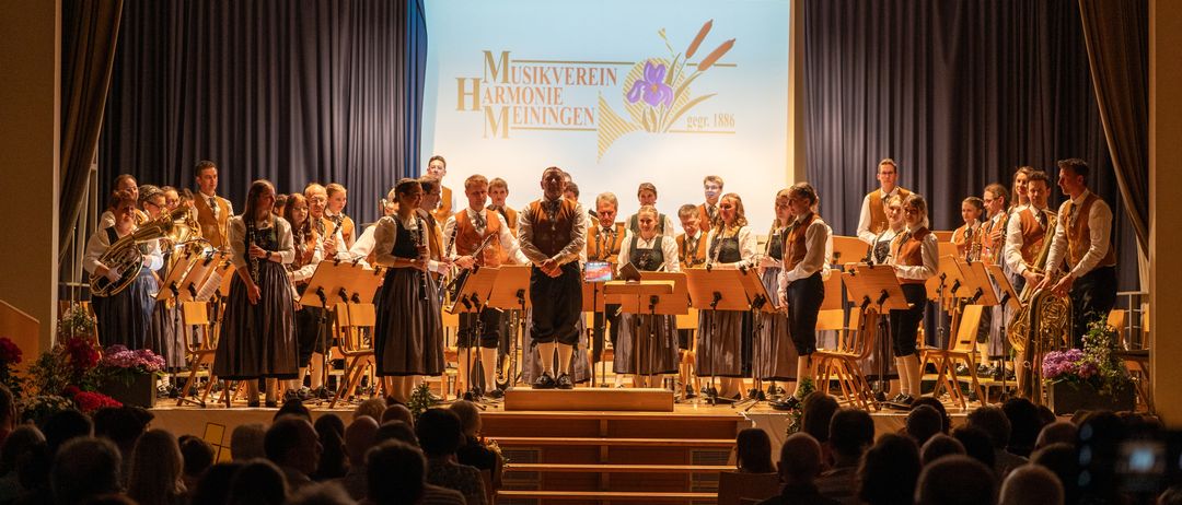 Ein Chor tritt auf der Bühne auf, begleitet vom Publikum. Auf der Bühne befinden sich Musikinstrumente und eine Projektionsfläche mit der Aufschrift 'Musikverein Harmonie Meiningen 1897-1886'.