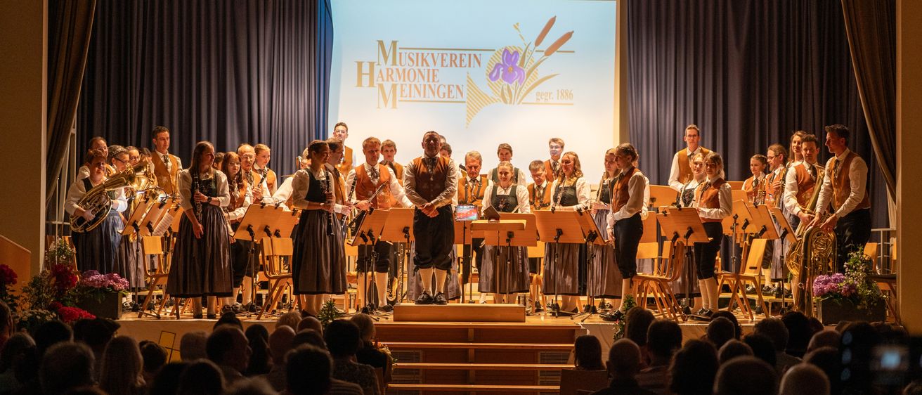 Ein Chor tritt auf der Bühne auf, begleitet vom Publikum. Auf der Bühne befinden sich Musikinstrumente und eine Projektionsfläche mit der Aufschrift 'Musikverein Harmonie Meiningen 1897-1886'.