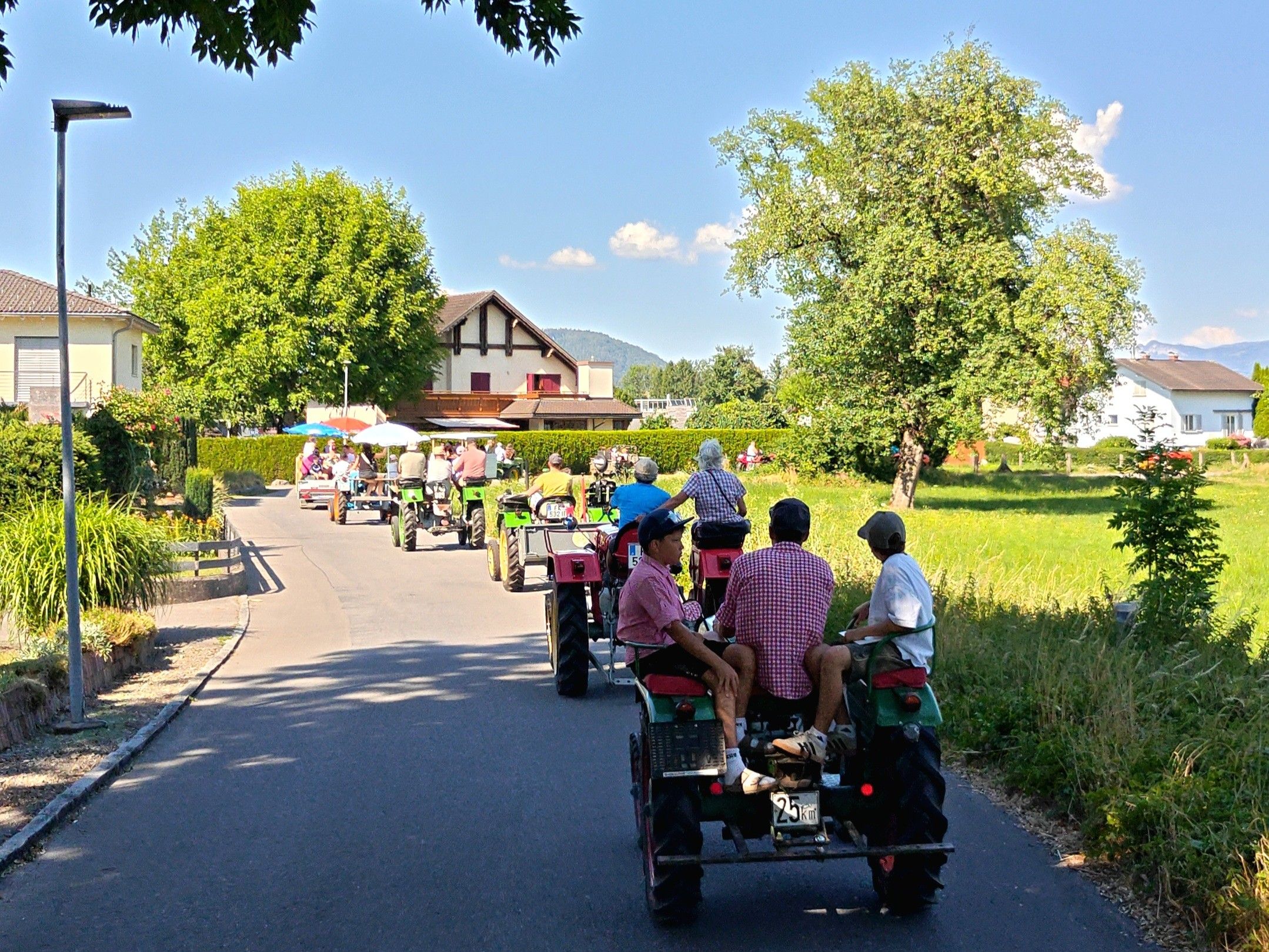 Eine Gruppe von Menschen fährt auf Traktoren eine Landstraße hinunter, mit einem Haus und Bergen im Hintergrund.