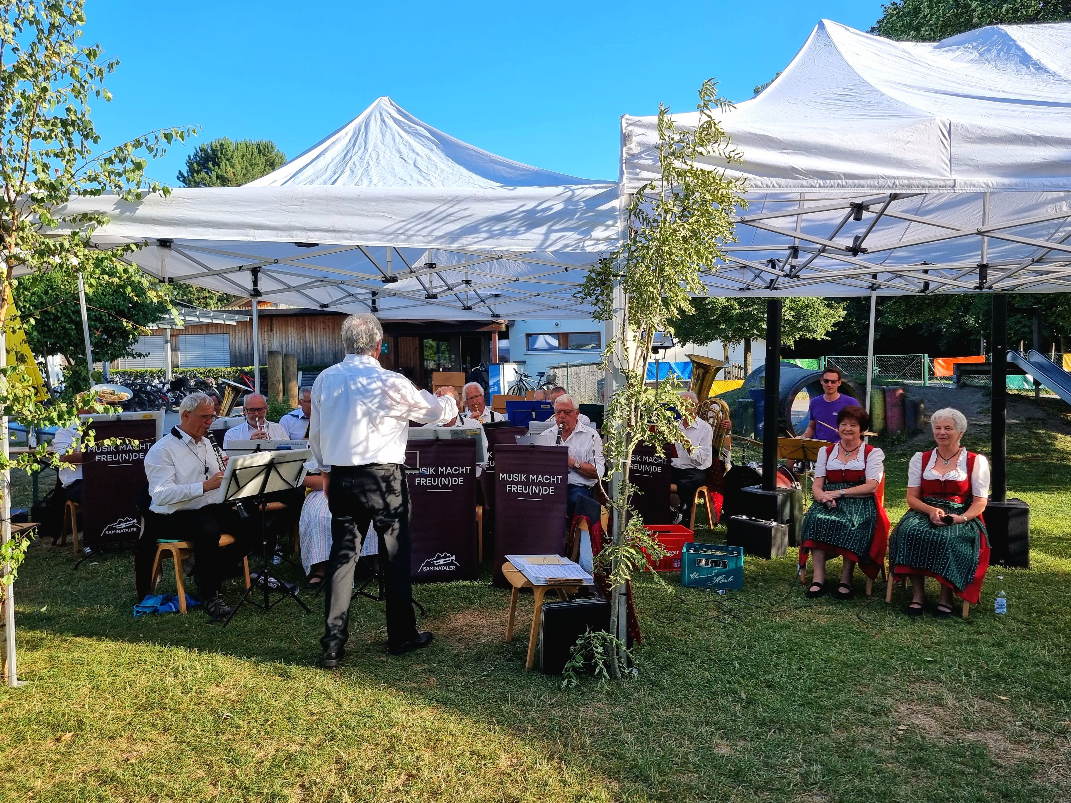 Eine Gruppe von Musikern spielt unter einem weißen Zelt mit einem Baum im Vordergrund. Einige Musiker sitzen auf Stühlen, während andere stehen. Eine Frau in einem traditionellen Kleid sitzt auf einem Stuhl rechts.