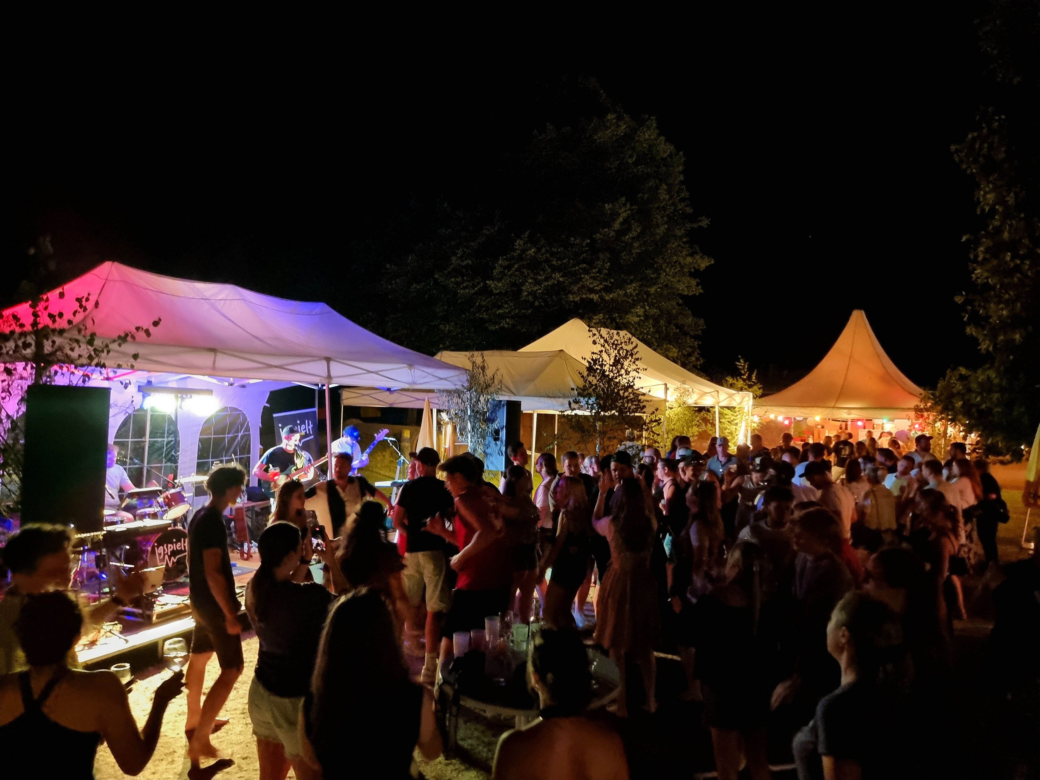 Eine nächtliche Veranstaltung im Freien, bei der Menschen um Zelte herum versammelt sind, einige tanzen, während andere eine Band auf der Bühne beobachten.