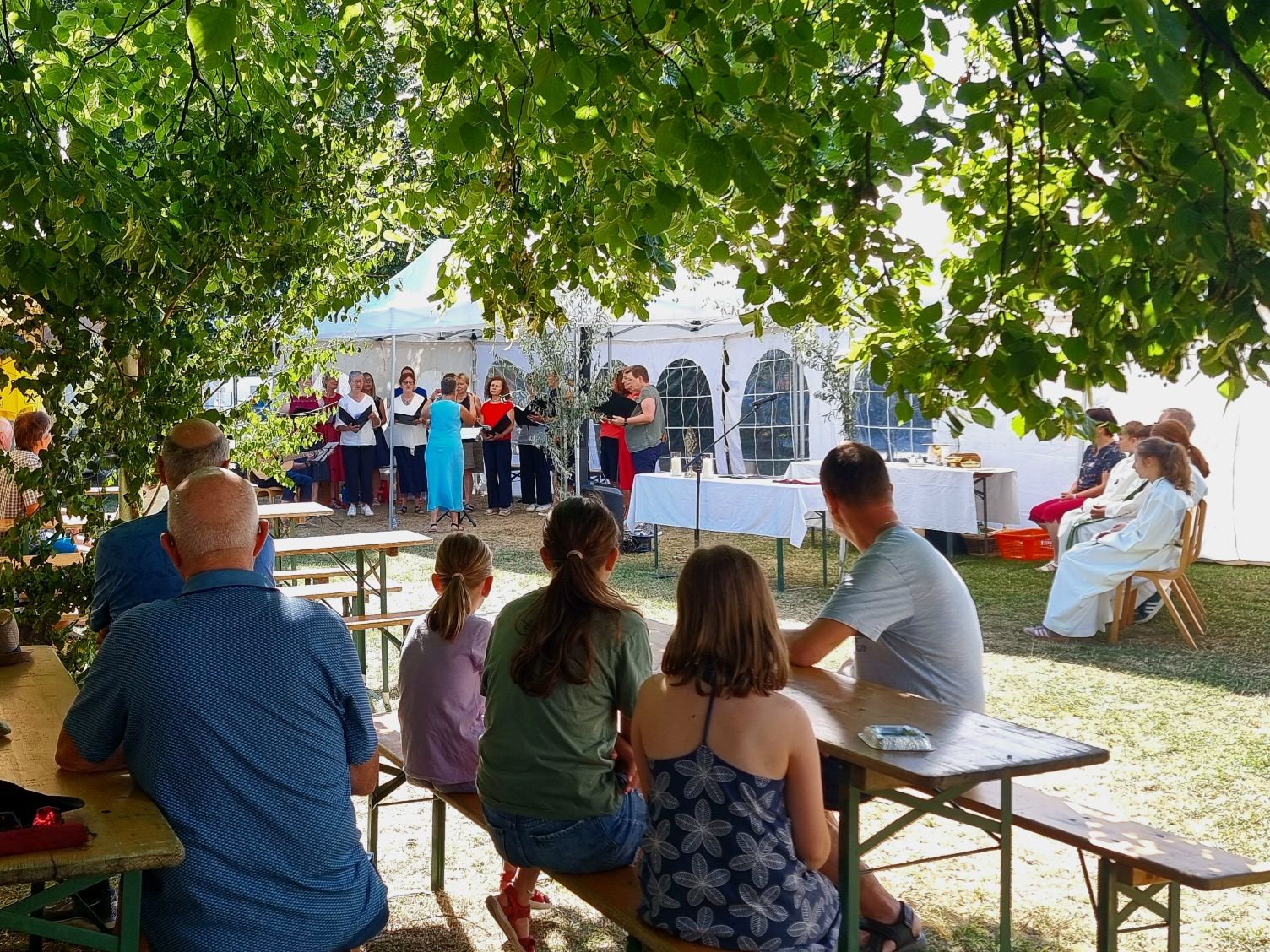 Eine Gruppe von Menschen versammelt sich im Freien unter einem Baum für eine Aufführung, einige sitzen auf Bänken und andere stehen und singen. Es gibt Tische mit Gegenständen darauf und Zelte im Hintergrund.