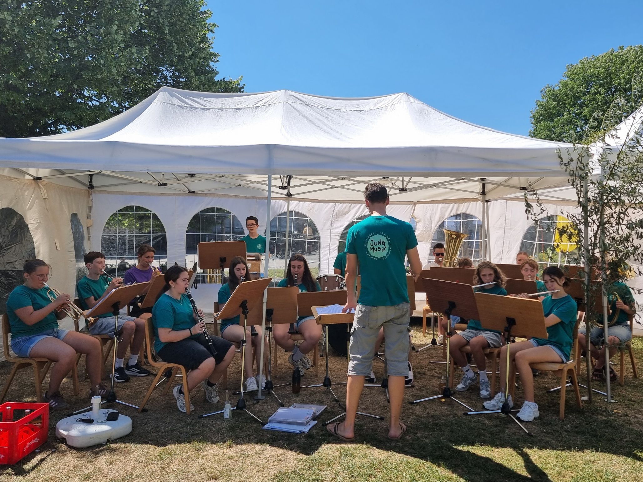 Eine Gruppe junger Musiker, in Grün gekleidet, sitzt unter einem weißen Zelt und übt mit ihren Instrumenten. Ein Mann steht vor ihnen, möglicherweise ihr Lehrer.