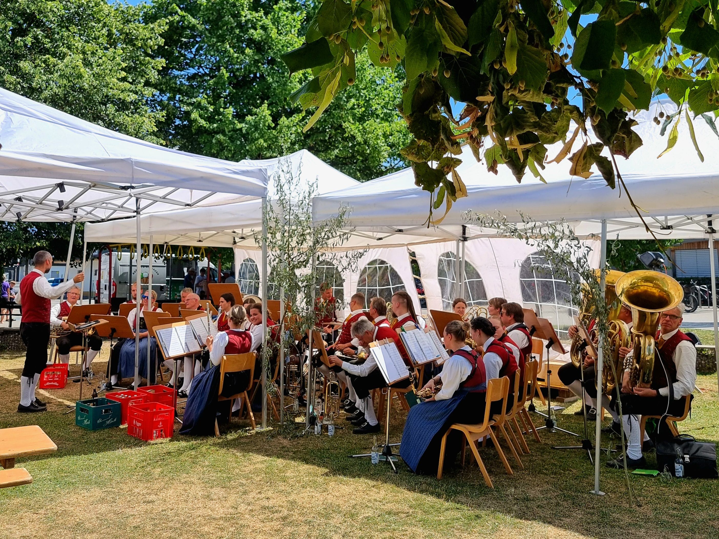 Ein Blasorchester spielt unter weißen Zelten im Freien, mit Musikern in traditioneller Kleidung und Instrumenten wie Tubas und Trompeten.