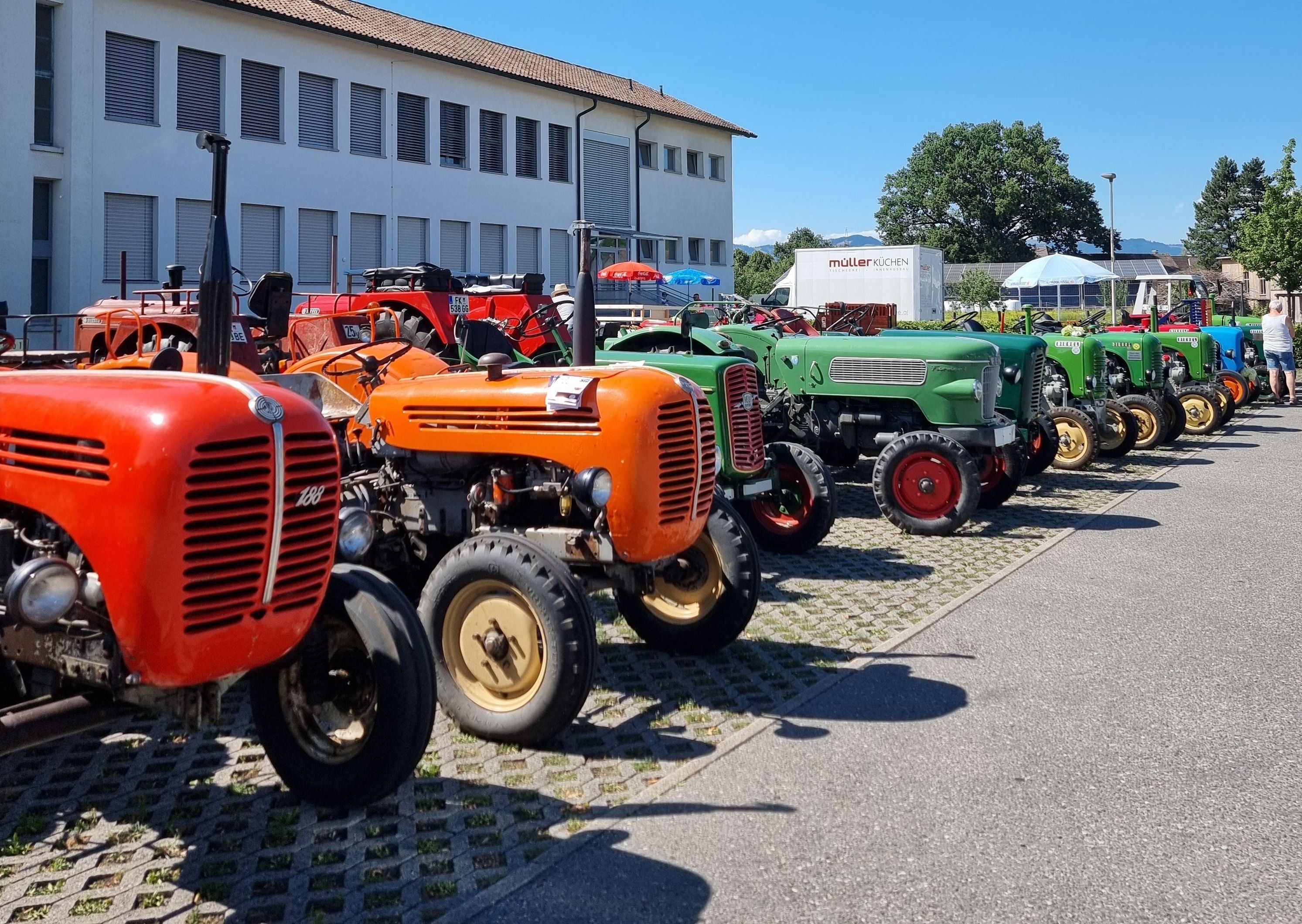 Eine Reihe von Oldtimer-Traktoren, darunter rote, orange und grüne Modelle, geparkt vor einem Gebäude mit weißer Wand und braunem Dach.
