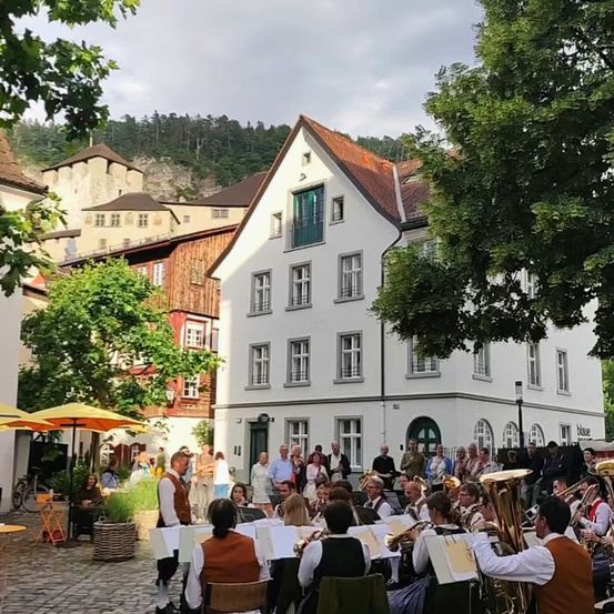 Ein Blasorchester spielt in einem offenen Innenhof vor einem großen weißen Gebäude. Das Publikum sitzt auf Stühlen und Bänken, und in der Nähe befinden sich Bäume und Schirme.