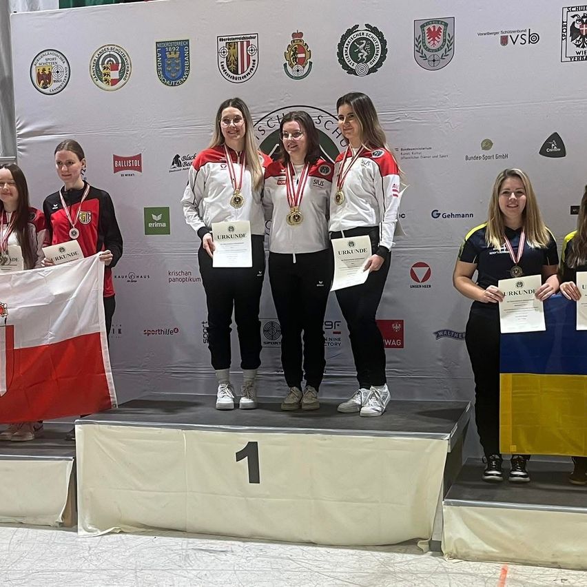Eine Gruppe von Athletinnen steht auf einem Podium, hält Medaillen und Urkunden in den Händen. Sie lächeln für die Kamera, eine hält eine polnische Flagge. Hinter ihnen ist eine Wand mit verschiedenen Logos.