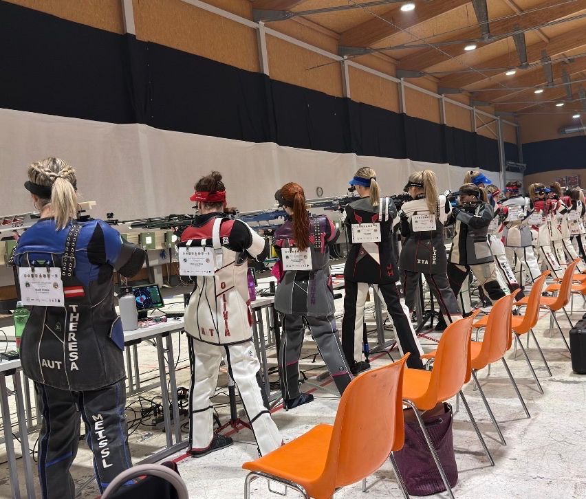 Eine Gruppe von Frauen mit Gewehren steht in einer überdachten Schießanlage in einer Reihe. Sie tragen Schießjacken und Stirnbänder. Einige von ihnen schauen sich etwas an. Orange Stühle sind vor ihnen angeordnet.