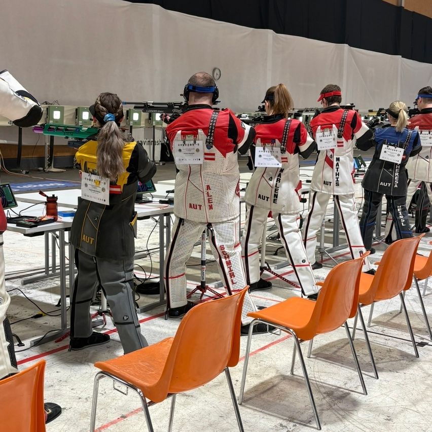 Eine Gruppe von Menschen nimmt an einem Schießwettbewerb teil. Sie tragen Uniformen mit roten und weißen Streifen und stehen in einer Reihe vor einer weißen Wand.