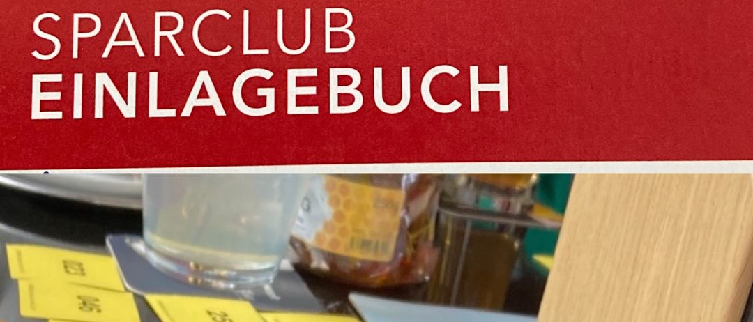 Eine rote Box mit der Aufschrift 'Sparclub Einlagebuch', gelben nummerierten Aufklebern, einem Glas und einem Papierbeutel auf einem Tisch.