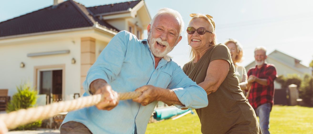 Eine Gruppe älterer Erwachsener spielt auf dem Rasen vor einem Haus Tauziehen. Sie alle lächeln und scheinen sich zu amüsieren.
