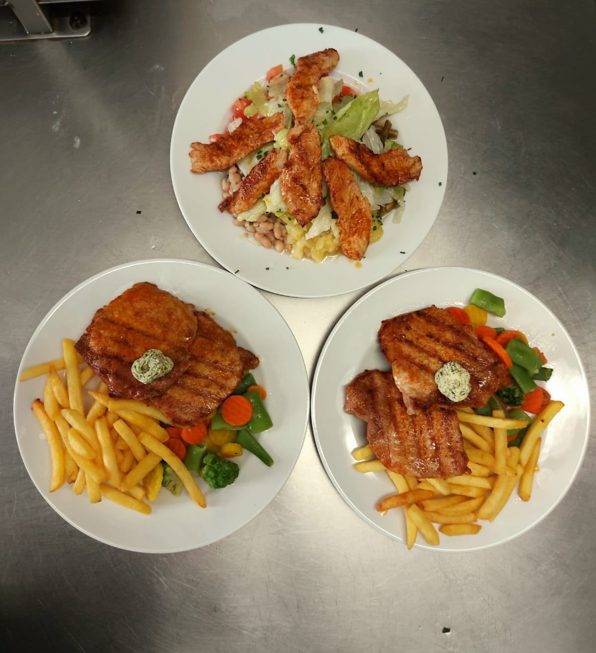 Drei Teller mit Essen auf einer Metalloberfläche. Ein Teller hat gegrilltes Hühnchen mit Salat. Ein anderer Teller hat Steak mit Pommes und Gemüse. Der dritte Teller hat gegrilltes Fleisch mit Pommes und Gemüse.