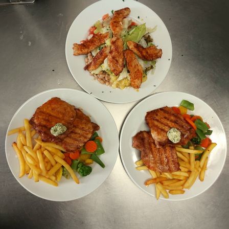 Drei Teller mit Essen auf einer Metalloberfläche. Ein Teller hat gegrilltes Hühnchen mit Salat. Ein anderer Teller hat Steak mit Pommes und Gemüse. Der dritte Teller hat gegrilltes Fleisch mit Pommes und Gemüse.