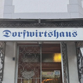 Ein Schild für Dorfwirtschaftshaus hängt über einem Ladeneingang. Das Schild ist weiß mit blauem Text. Eine Lampe beleuchtet den Eingang. Eine Glastür ist sichtbar, mit einer Lichtreflexion im Inneren.