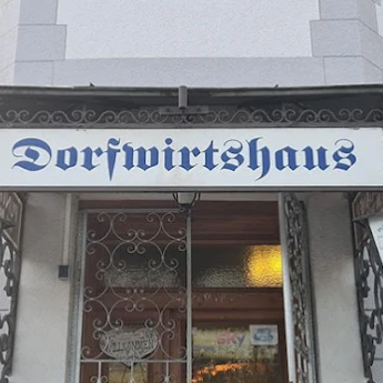 Ein Schild für Dorfwirtschaftshaus hängt über einem Ladeneingang. Das Schild ist weiß mit blauem Text. Eine Lampe beleuchtet den Eingang. Eine Glastür ist sichtbar, mit einer Lichtreflexion im Inneren.