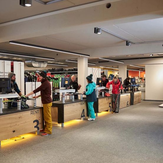 Ein Skiverleih mit mehreren Kunden am Tresen und Mitarbeitern dahinter. Der Laden ist gut beleuchtet mit Deckenleuchten.
