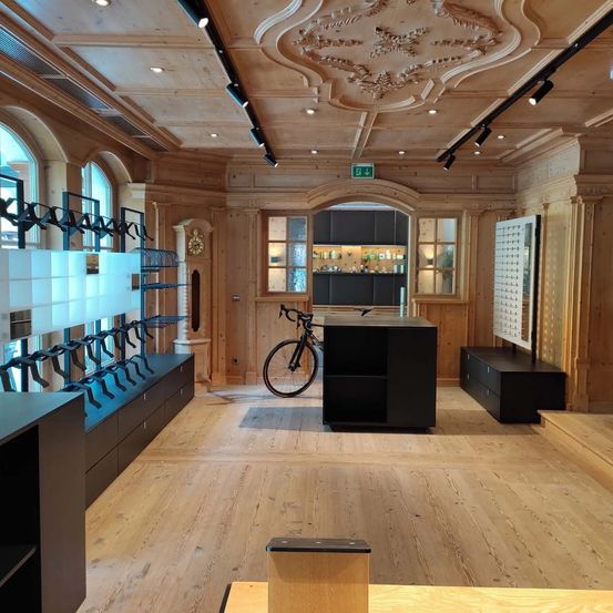 Innenansicht eines modernen Geschäfts mit hölzernen Wänden, Böden und Regalen. Ein Fahrrad steht in der Mitte, und Regale mit Fahrradteilen befinden sich links.