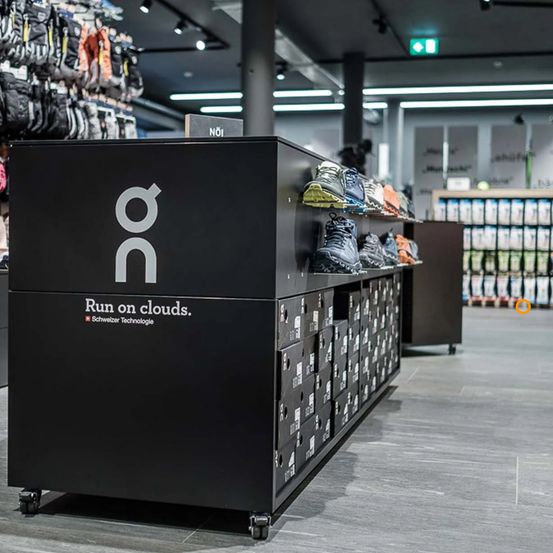 Ein Schuhdisplay in einem Geschäft mit dem Text 'Laufe auf Wolken' und Schweizer Technologie, mit Schuhen auf den Regalen, Aufbewahrungsboxen und einem Rucksack im Hintergrund.