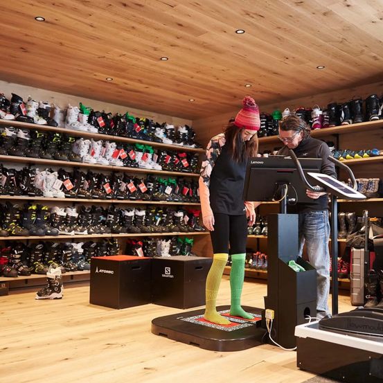 Eine Frau mit Hut und bunten Socken steht auf einer Waage mit einem Mann in einer Brille. Sie betrachten einen Monitor in einem Geschäft mit Regalen voller Skischuhe.