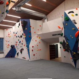Innenansicht eines Kletterzentrums mit bunten Klettergriffen und einem breiten grauen Boden.