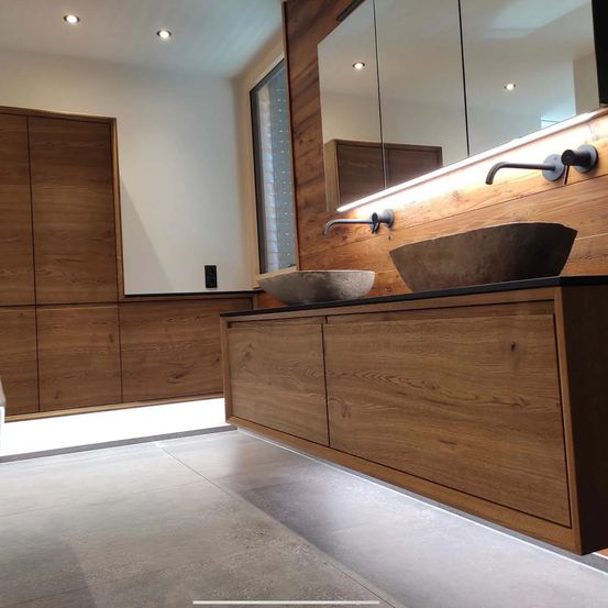 Modernes Badezimmer mit Holzschrank und zwei Steinwaschbecken auf einer Holzarbeitsplatte. Zwei Wasserhähne und zwei Spiegel mit Beleuchtung sind an der Holzwand angebracht.