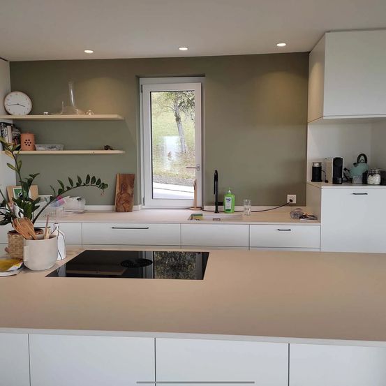 Eine moderne Küche mit einem beigen Arbeitstisch und weißen Schränken. Regale an der Wand halten eine Uhr, Bücher und eine Vase. Ein Fenster lässt natürliches Licht eindringen, und eine Tür führt nach draußen.