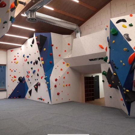 Ein Indoor-Kletterzentrum mit weißen Wänden und verschiedenen farbigen Klettergriffen, einem grauen Boden und einer großen weißen Wand mit einem gelben Kreis darauf.