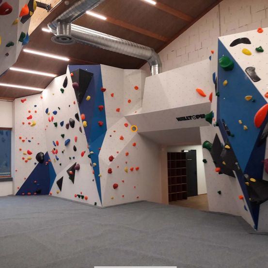 Ein Indoor-Kletterzentrum mit weißen Wänden und verschiedenen farbigen Klettergriffen, einem grauen Boden und einer großen weißen Wand mit einem gelben Kreis darauf.