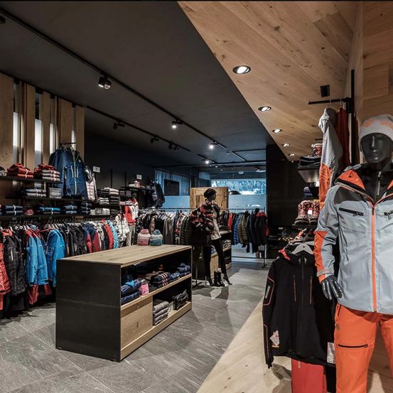 Ein Bekleidungsgeschäft mit Holzdecke und gefliestem Boden. Verschiedene Kleidungsstücke hängen an den Kleiderstangen und einem Holzregal. Ein Mannequin ist in orangefarbener Hose und einer grauen Jacke mit einer weißen Mütze gekleidet.